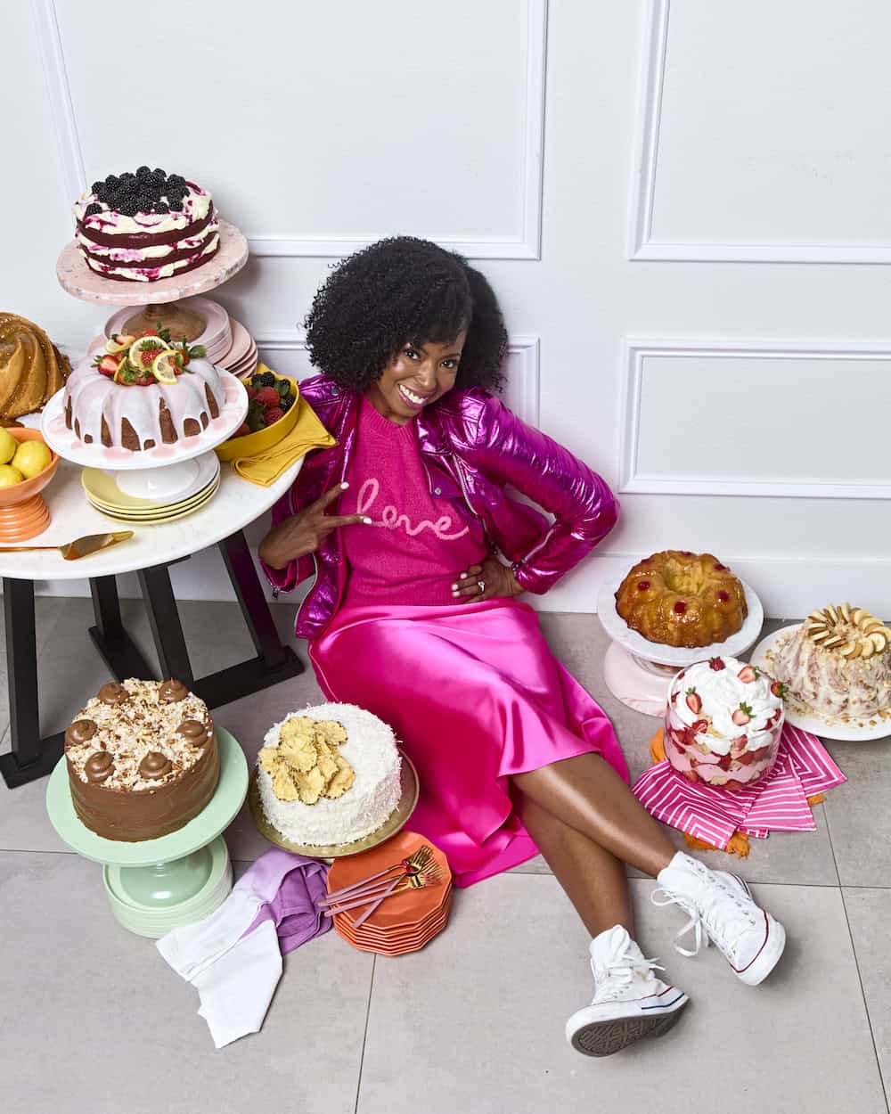 Jocelyn Delk Adams smiling while sitting with amazing pound cakes and layer cakes surrounding her