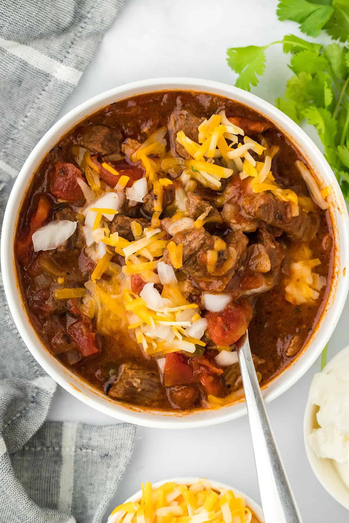 Overhead shot of a bowl of brisket chili topped with shredded cheese, sour cream, and cilantro, with a kitchen cloth and a bunch of cilantro next to it