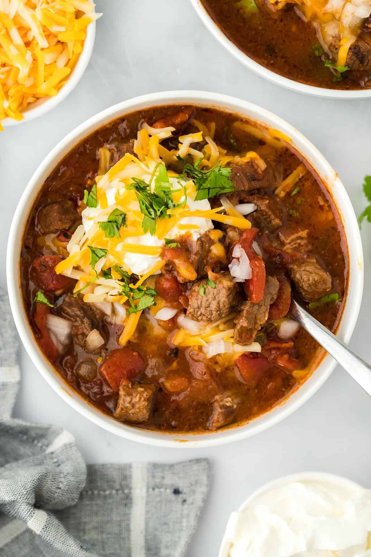 Overhead shot of a bowl of brisket chili topped with shredded cheese, sour cream, and cilantro, with more chili, a bowl of shredded cheese, and a bowl of sour cream next to it