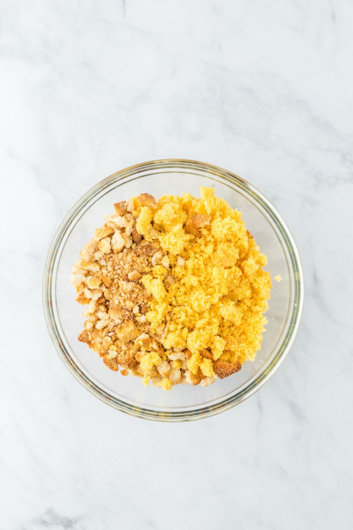 A clear glass mixing bowl with crumbled cornbread and crushed croutons