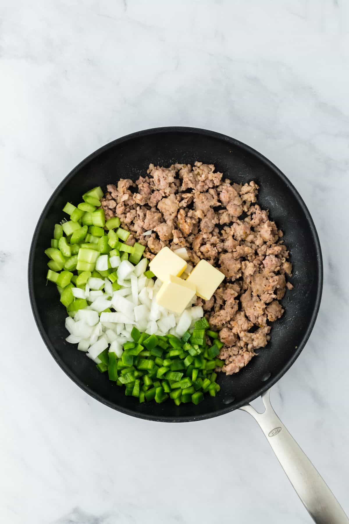 Cooked sausage with chopped green bell peppers, onions, celery, and pats of butter in the skillet