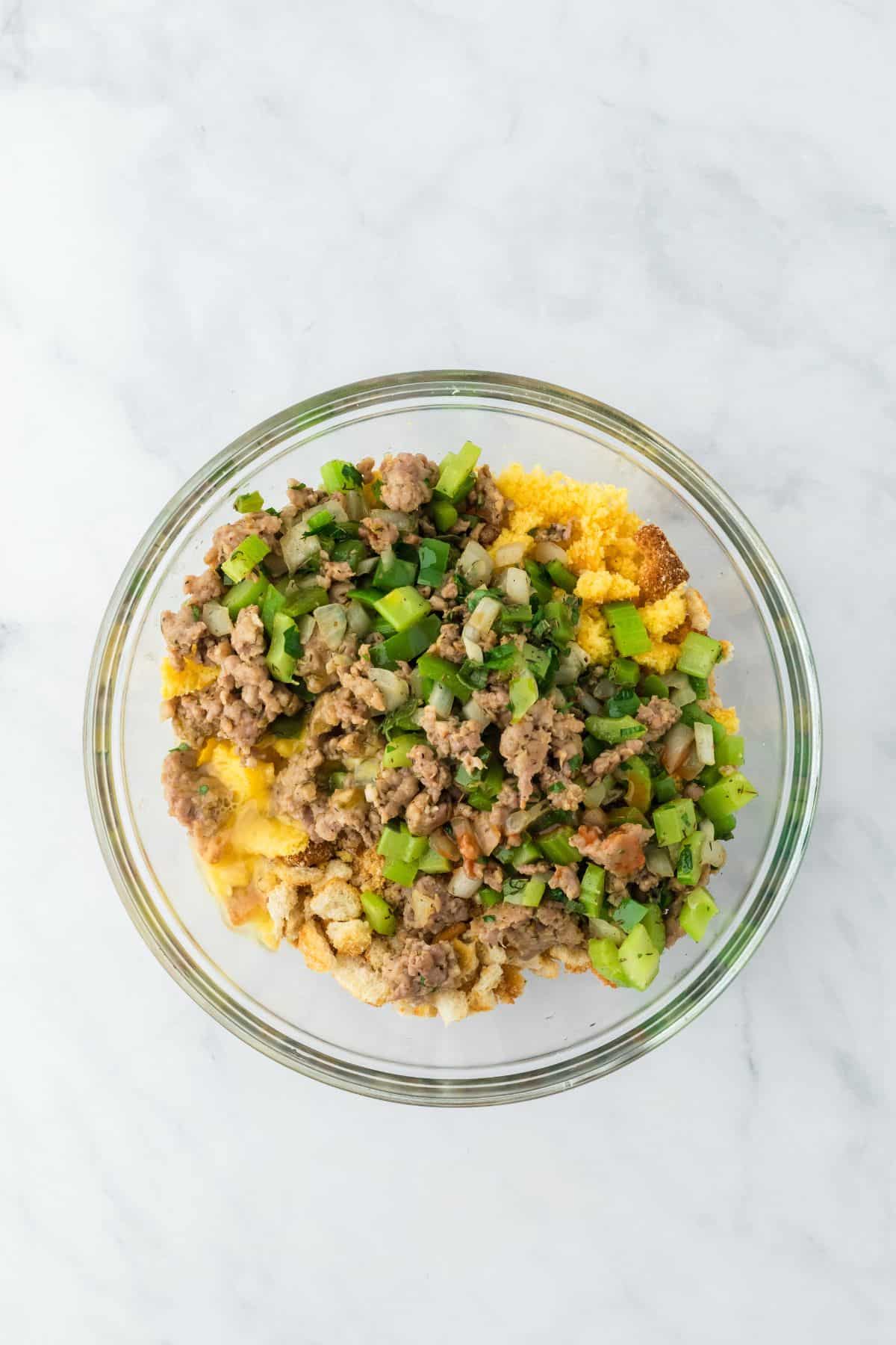 A large mixing bowl with cornbread crumbs and crouton crumbs, topped with cooked sausage and vegetable mixture