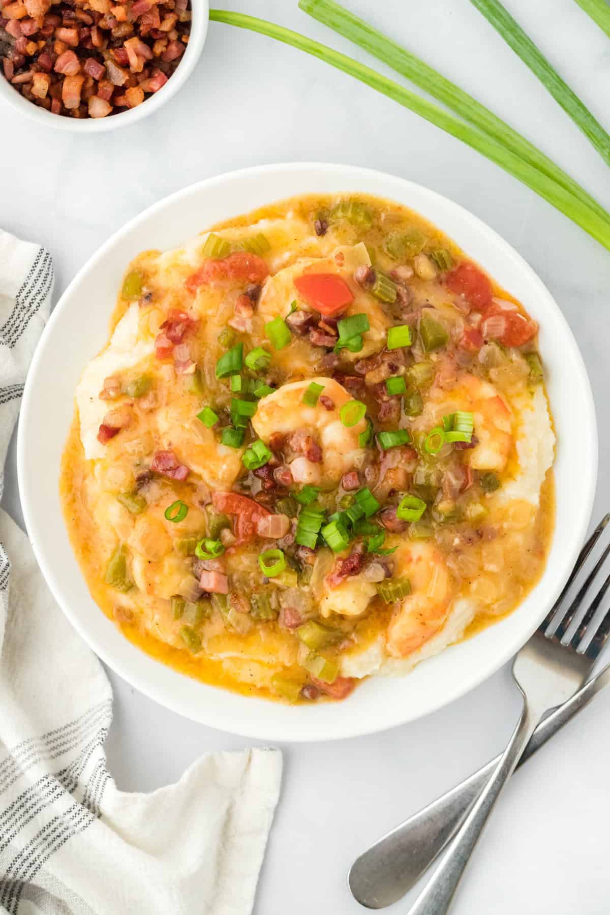 Grits and shrimp plated on a white plate with green onions and pancetta in the white background with two silver forks