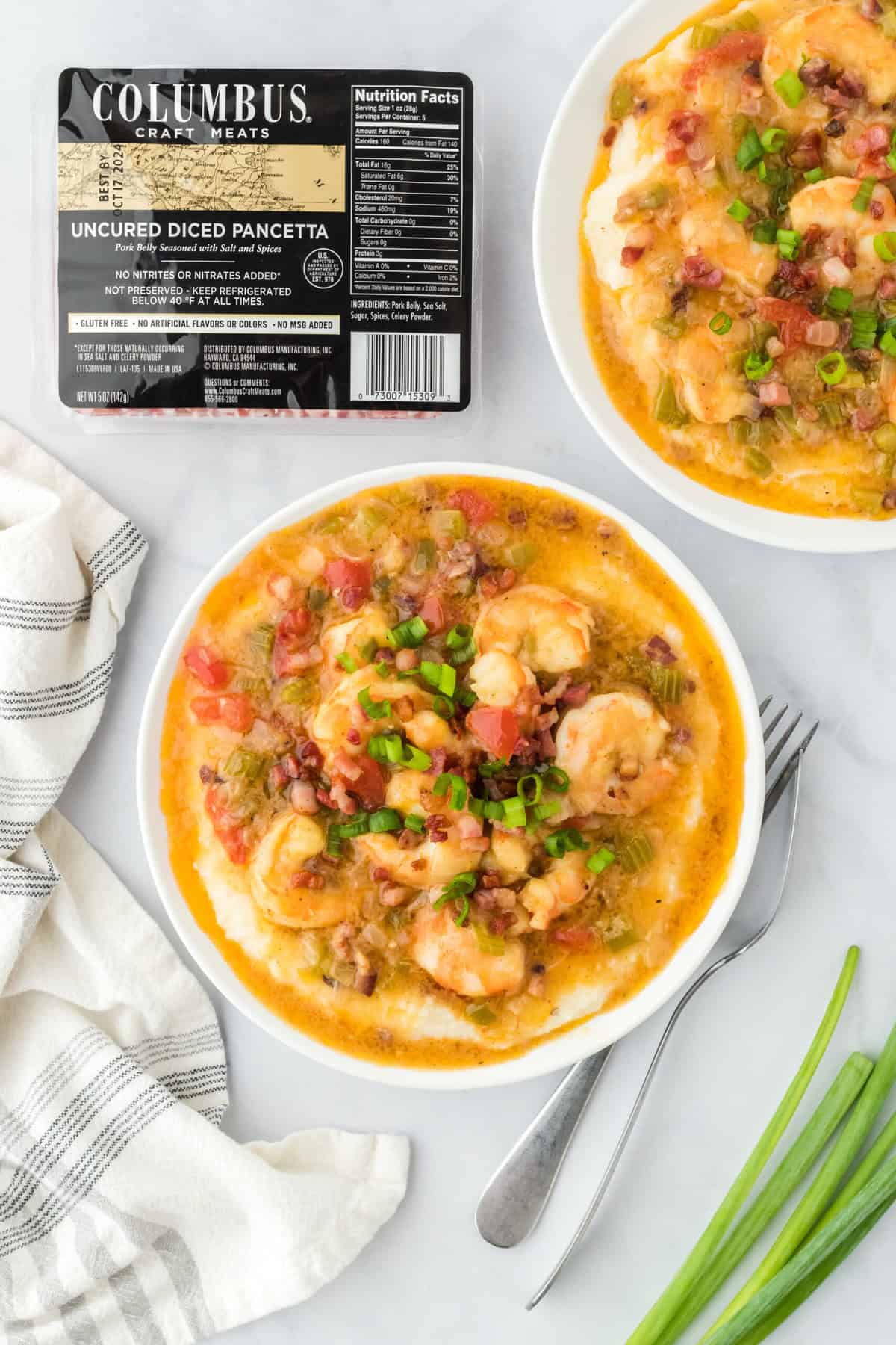 Two bowls of New Orleans shrimp and grits on white countertop with pancetta package in background