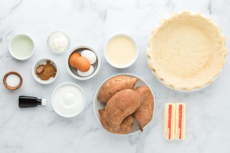 Sweet potatoes, pie dough, butter, spices, sugar in white bowls on white countertop to make a pie