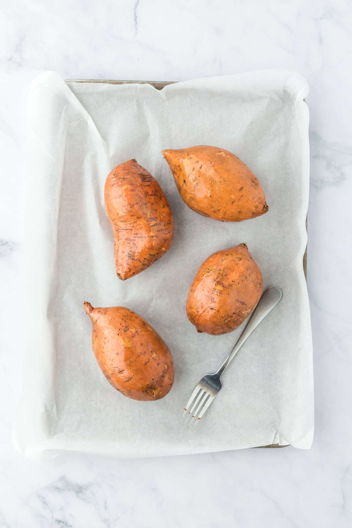 Four sweet potatoes rubbed in oil on parchment baking sheet on white countertop