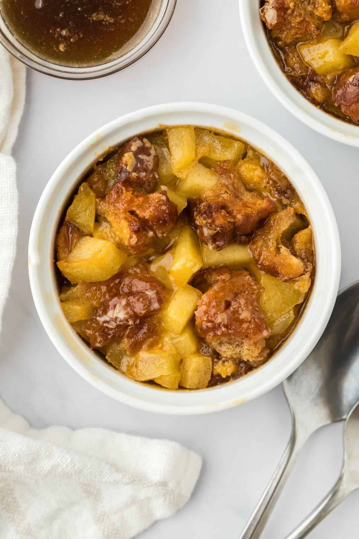 Overhead shot of a ramekin filled with golden-brown apple fritter bread pudding with more ramekins and a couple of spoons next to it.