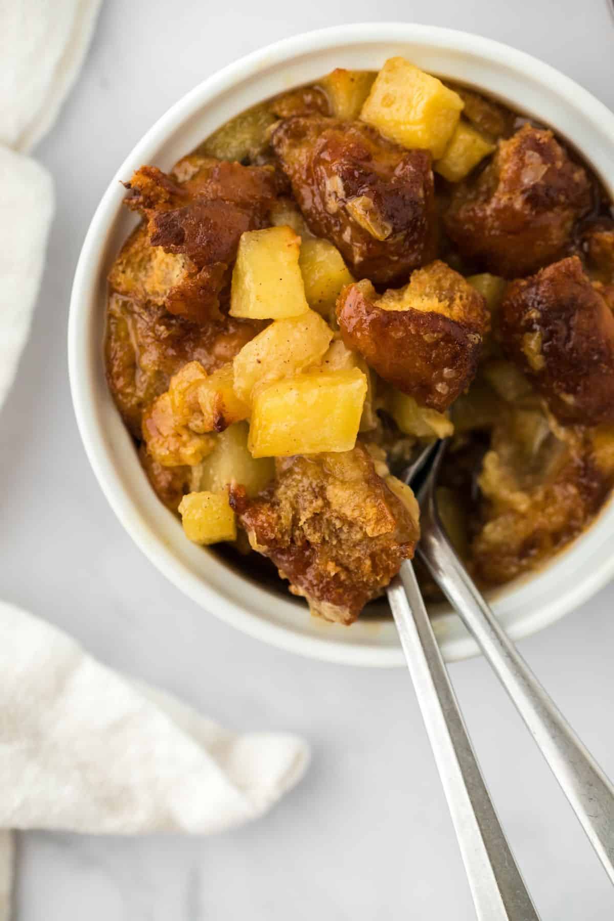 Overhead shot of a ramekin filled with golden-brown apple fritter bread pudding with two spoons in it.