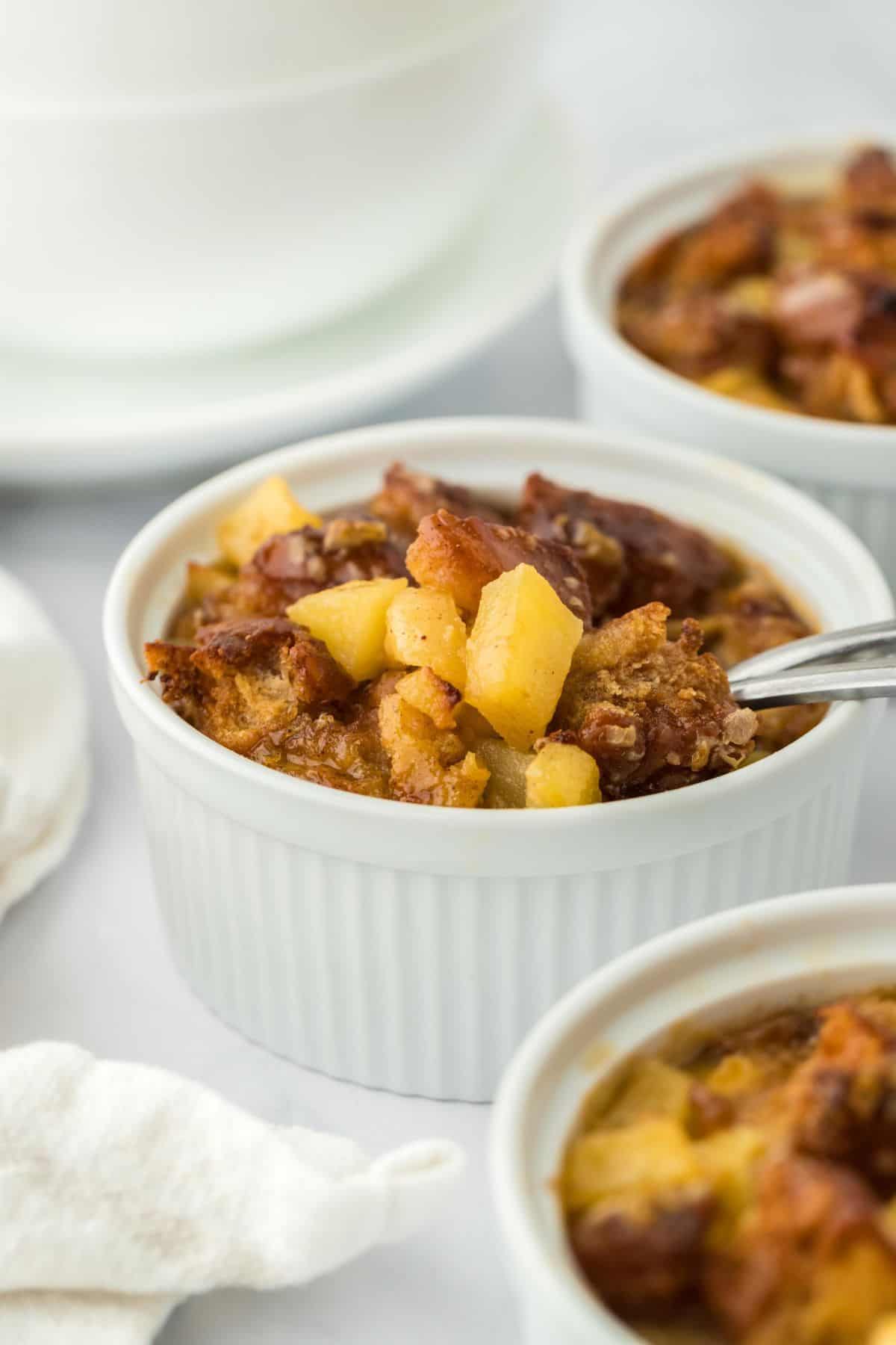 Closeup of a small ramekin filled with golden-brown apple fritter bread pudding with more ramekins in the background.