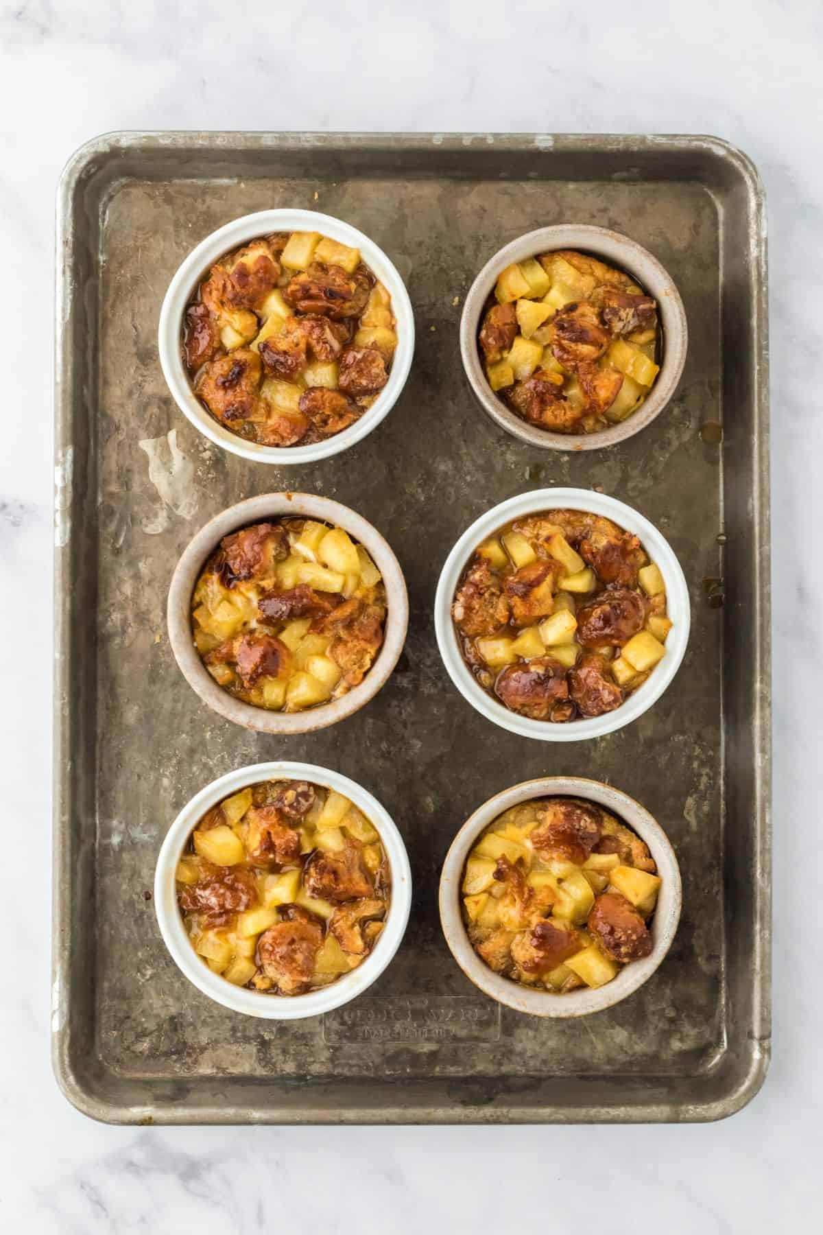 Six ramekins filled with baked apple fritter bread pudding, golden and slightly caramelized.