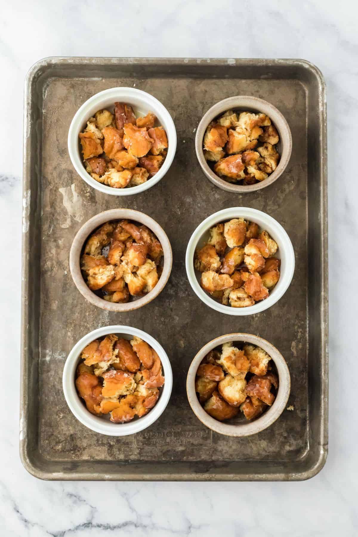 Ramekins on a baking tray filled with torn bread pieces, prepped for the bread pudding base.