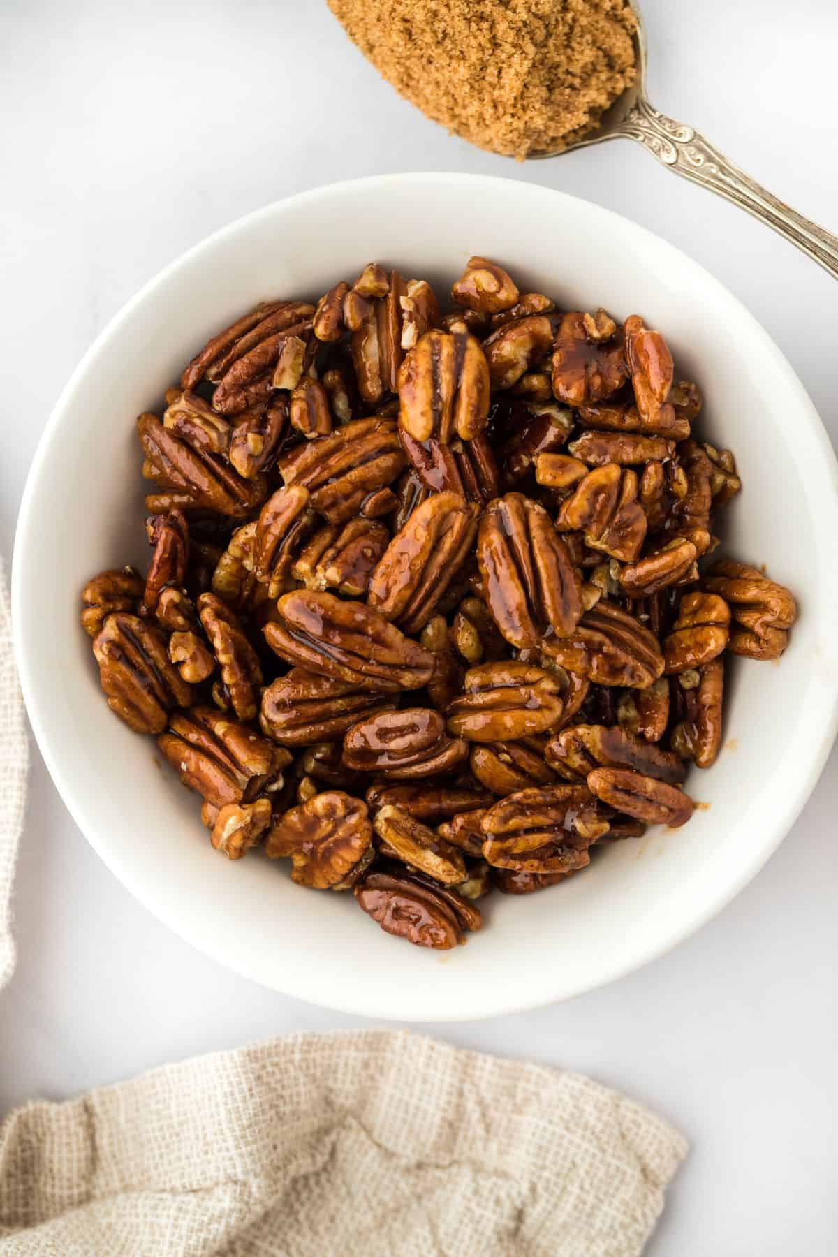 Overhead shot of a bowl full of candied pecans next to a spoonful of brown sugar