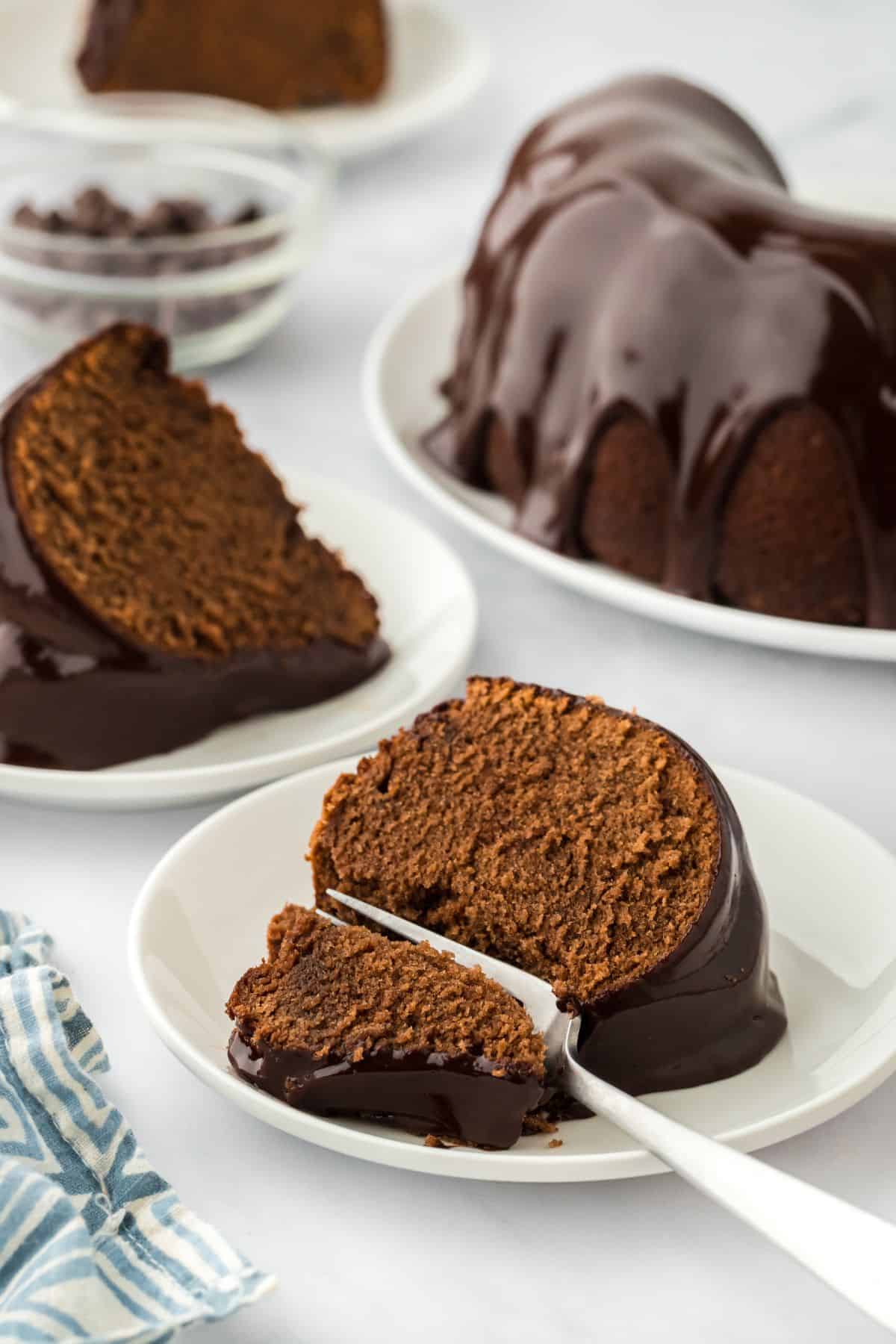 A slice of chocolate pound cake drizzled with chocolate glaze on a white plate, with more cake slices and a whole bundt cake in the background