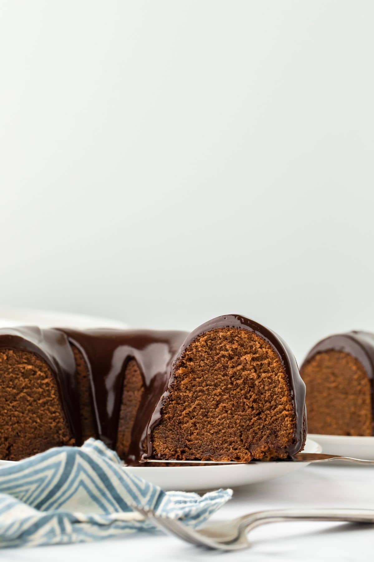 A side view of a cut chocolate pound cake with a smooth chocolate glaze dripping down, with a blue napkin in the foreground and more cake in the background