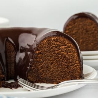 A side view of a cut chocolate pound cake with a smooth chocolate glaze dripping down, with a blue napkin in the foreground and more cake in the background