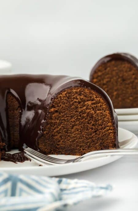 A side view of a cut chocolate pound cake with a smooth chocolate glaze dripping down, with a blue napkin in the foreground and more cake in the background