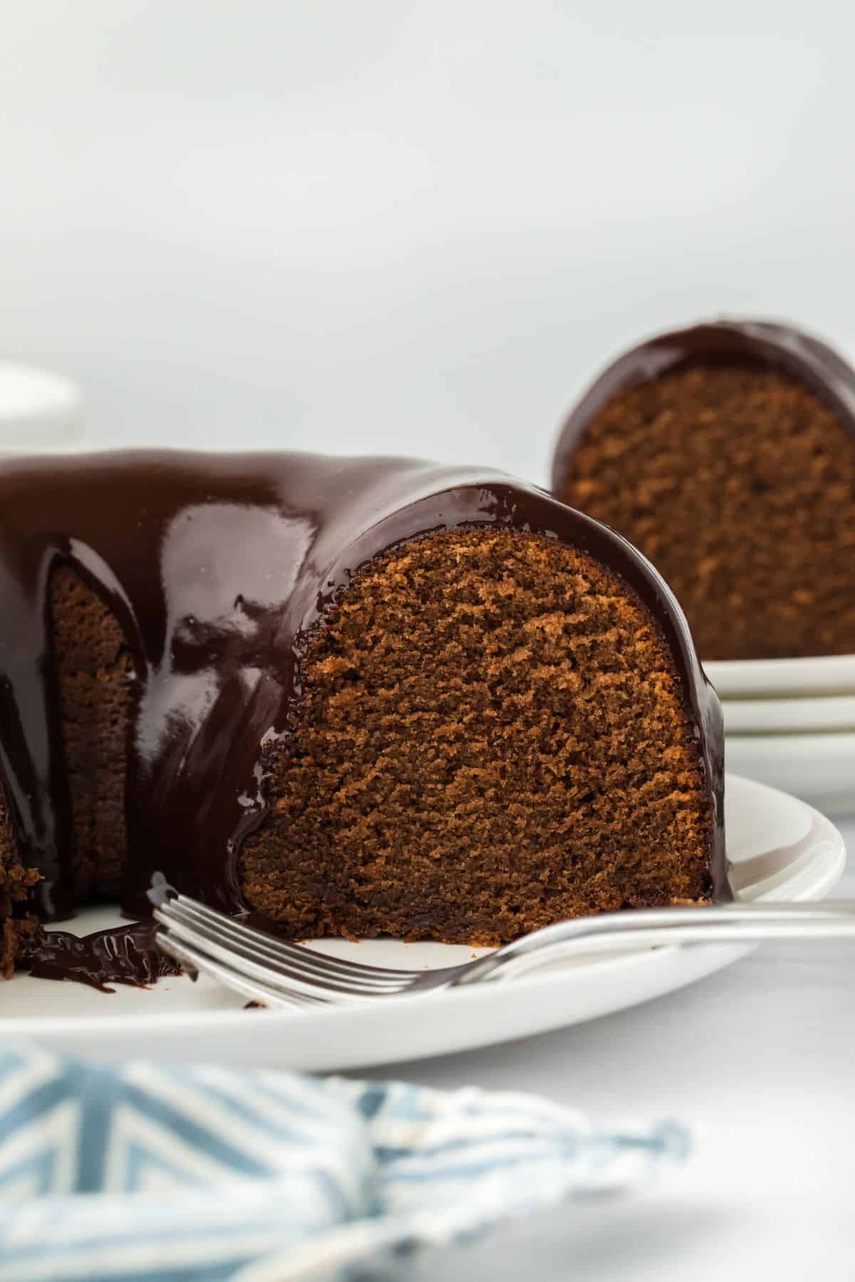 A side view of a cut chocolate pound cake with a smooth chocolate glaze dripping down, with a blue napkin in the foreground and more cake in the background