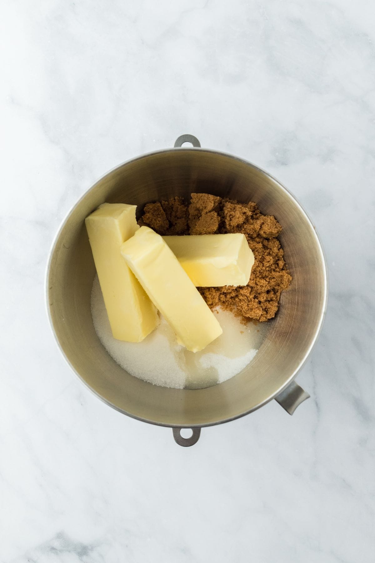 A metal mixing bowl filled with butter sticks, brown sugar, and white sugar