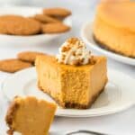 A slice of pumpkin cheesecake topped with whipped cream and crushed gingersnaps, with a fork cutting into it and more cookies in the background