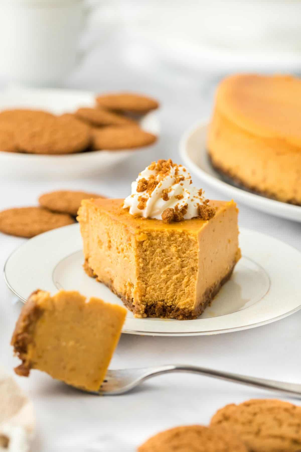 A slice of pumpkin cheesecake topped with whipped cream and crushed gingersnaps, with a fork cutting into it and more cookies in the background