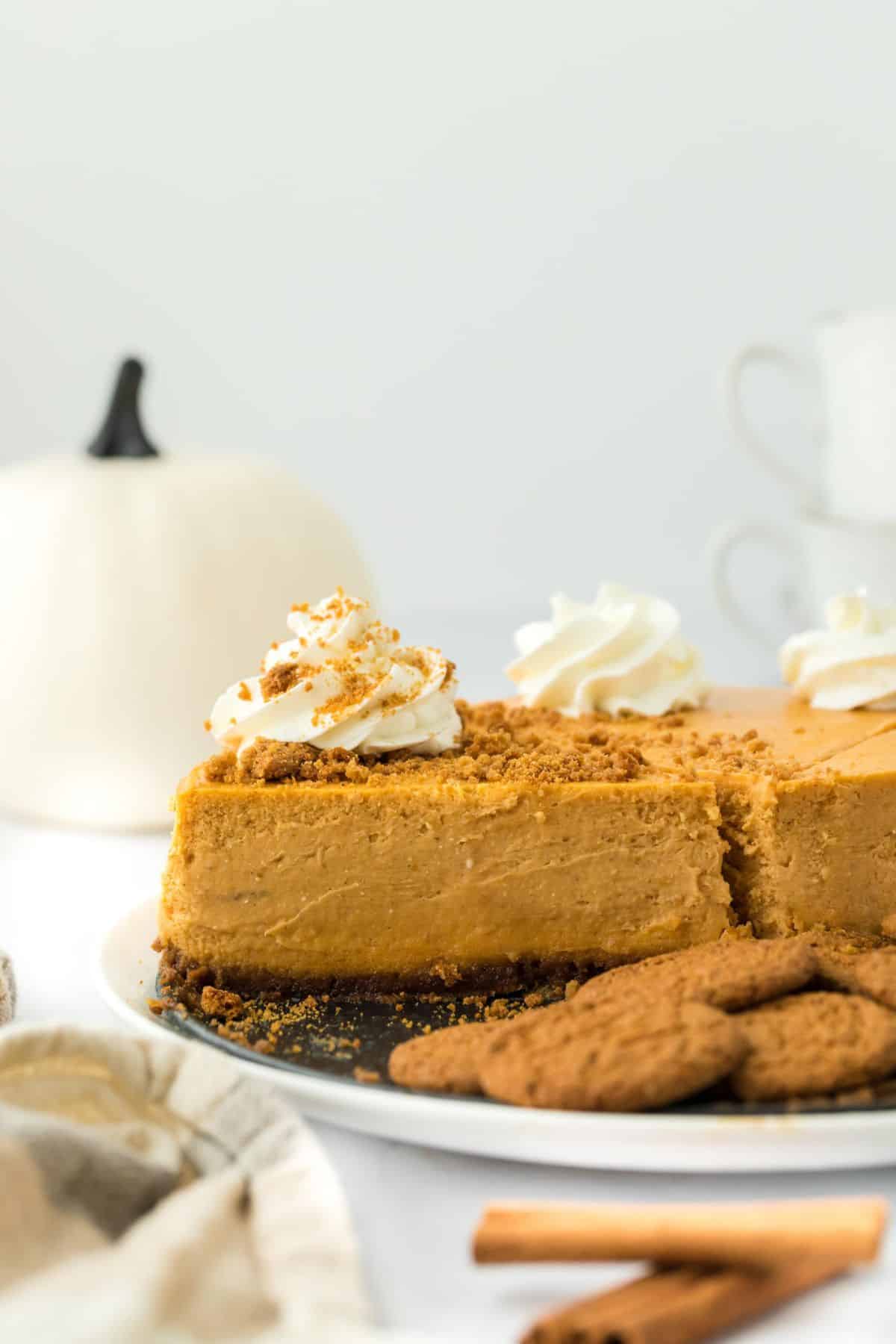 A sliced pumpkin cheesecake topped with whipped cream and crushed gingersnaps, with more cookies next to it and a white pumpkin in the background.