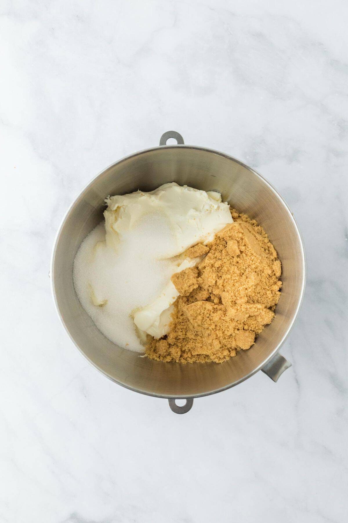 A mixing bowl with cream cheese, sour cream, heavy cream, brown sugar and granulated sugar piled together before mixing