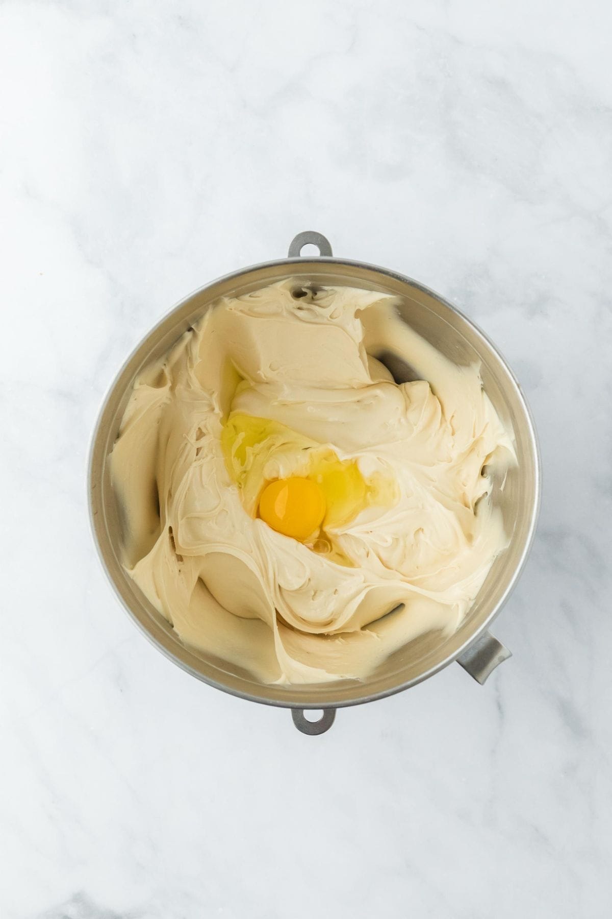 A smooth, creamy cheesecake mixture with an egg added on top, in a metal mixing bowl