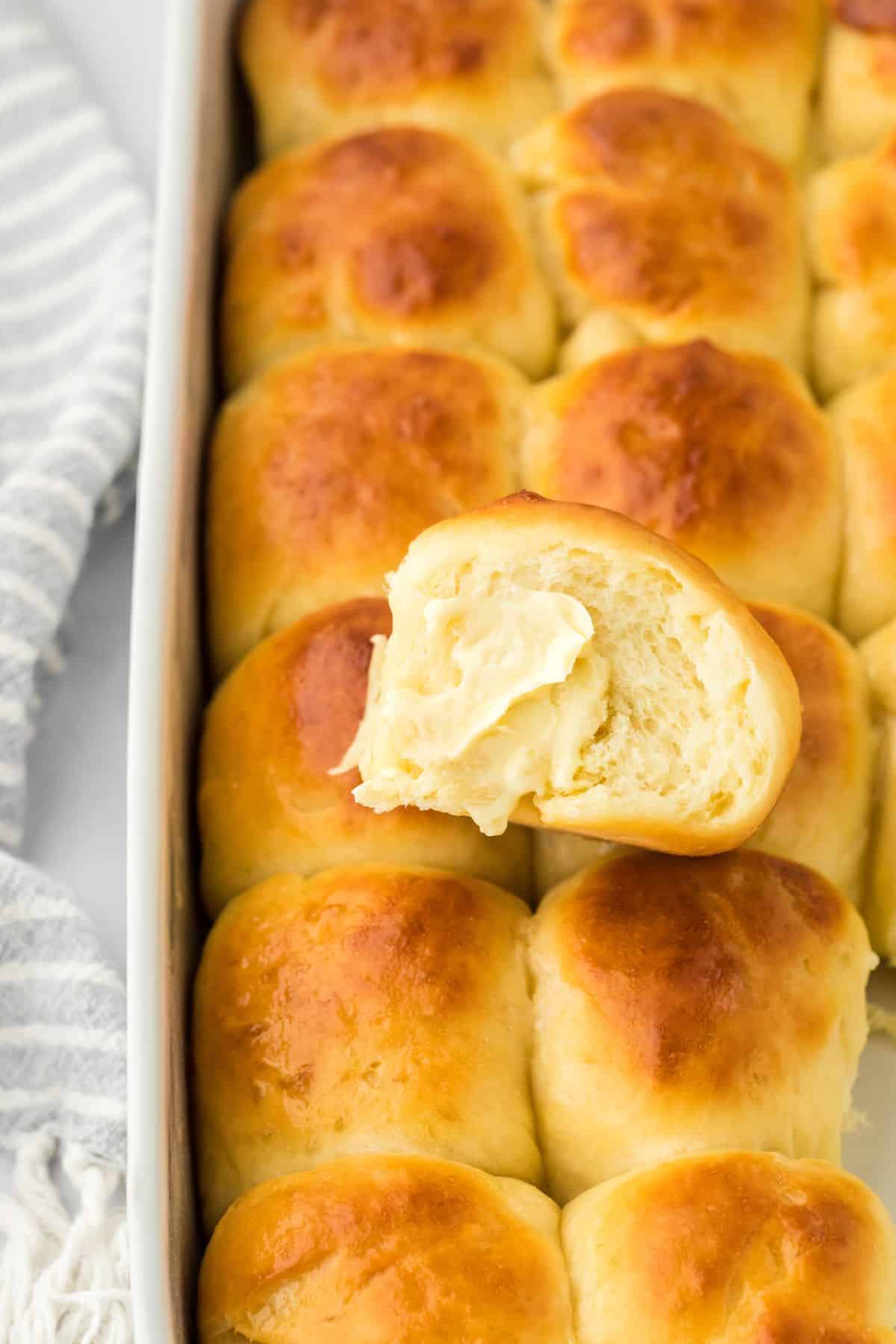 A single yeast roll with butter spread on top, resting on the edge of a baking dish filled with more golden rolls