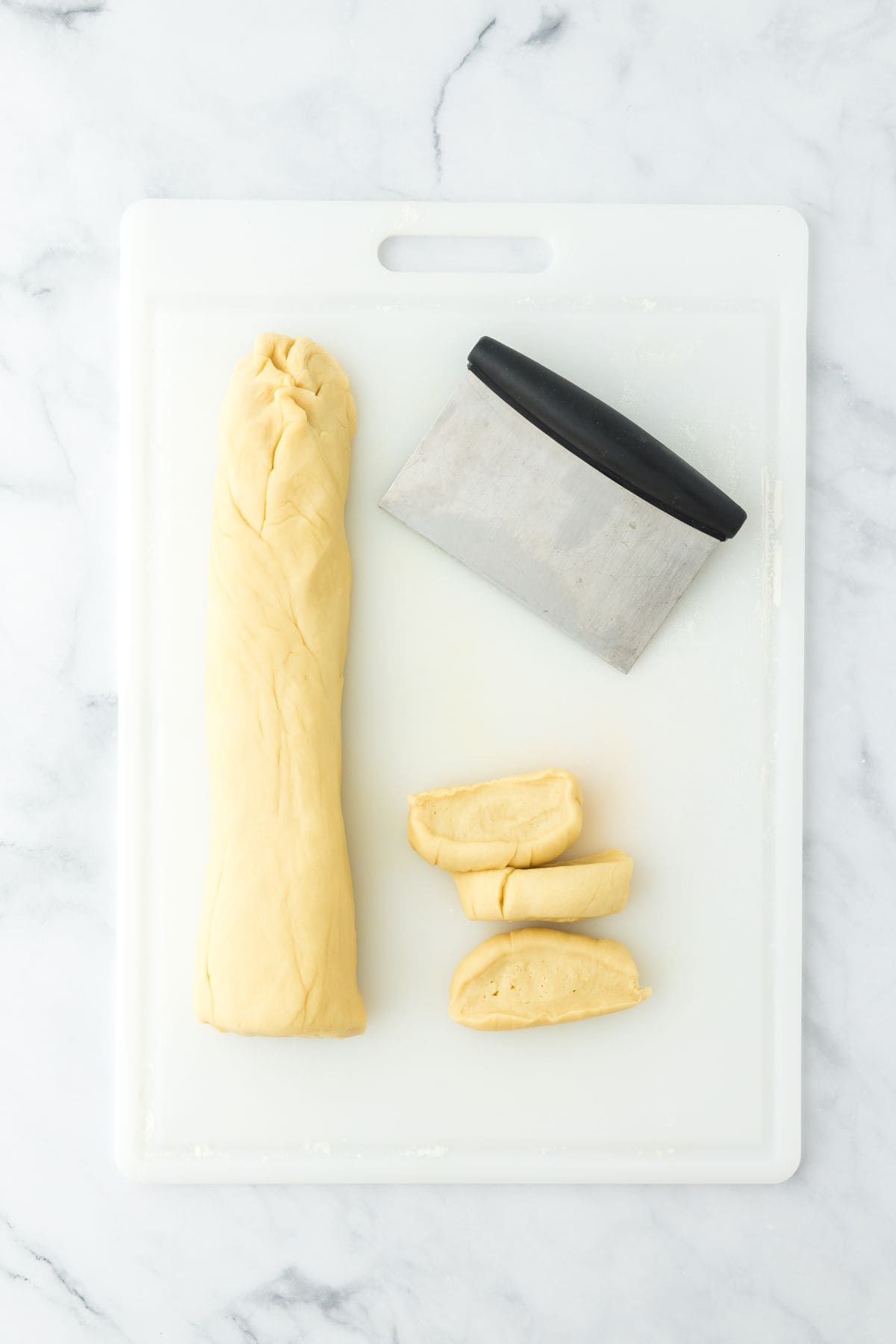 A long roll of dough and a dough scraper on a cutting board, with some dough pieces cut off