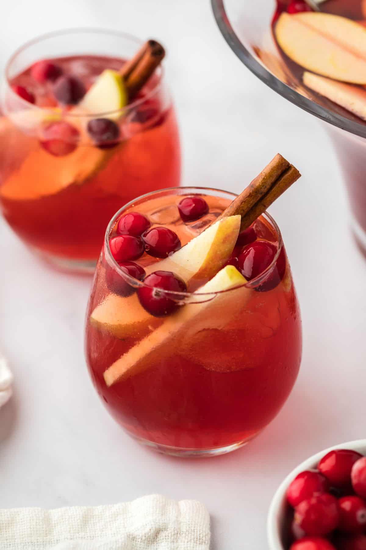 Two glasses of red Thanksgiving punch with ice, cranberries, and apple slices, garnished with a cinnamon stick