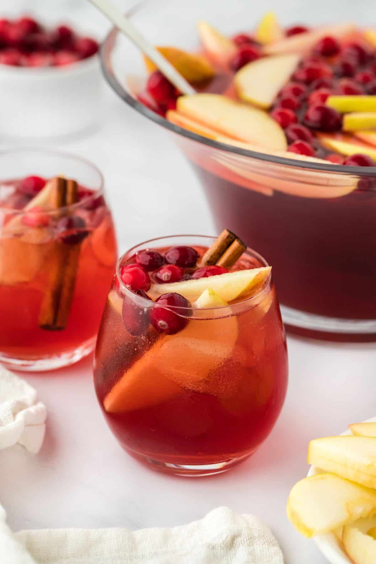 Two glasses of red Thanksgiving punch with ice, cranberries, and apple slices, garnished with a cinnamon stick, with a punch bowl in the background