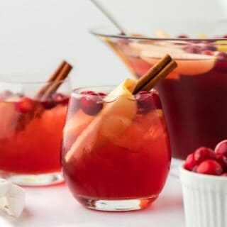 Two glasses of red Thanksgiving punch, garnished with a cinnamon stick and apple slices, with a punch bowl and ingredients in the background