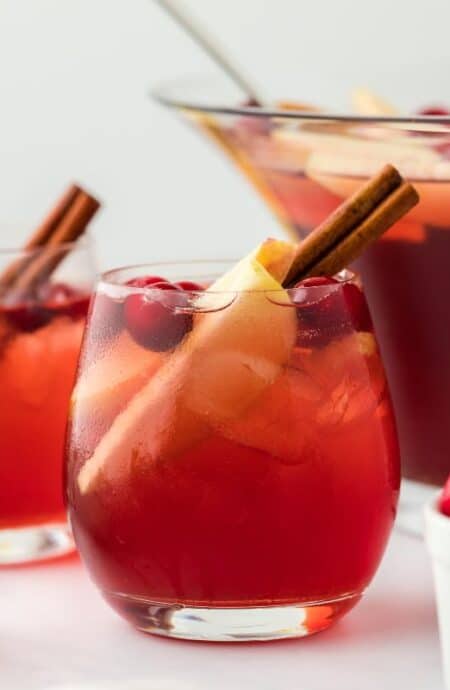 Two glasses of red Thanksgiving punch, garnished with a cinnamon stick and apple slices, with a punch bowl and ingredients in the background