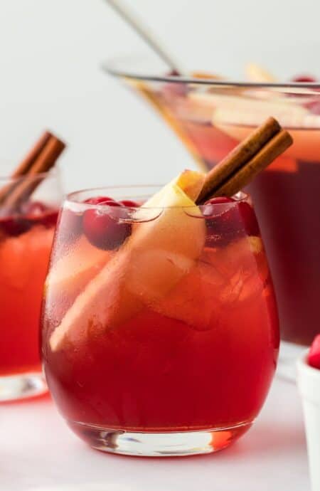 Close-up shot of a glass of red Thanksgiving punch, garnished with a cinnamon stick and apple slices, with punch bowl in the background