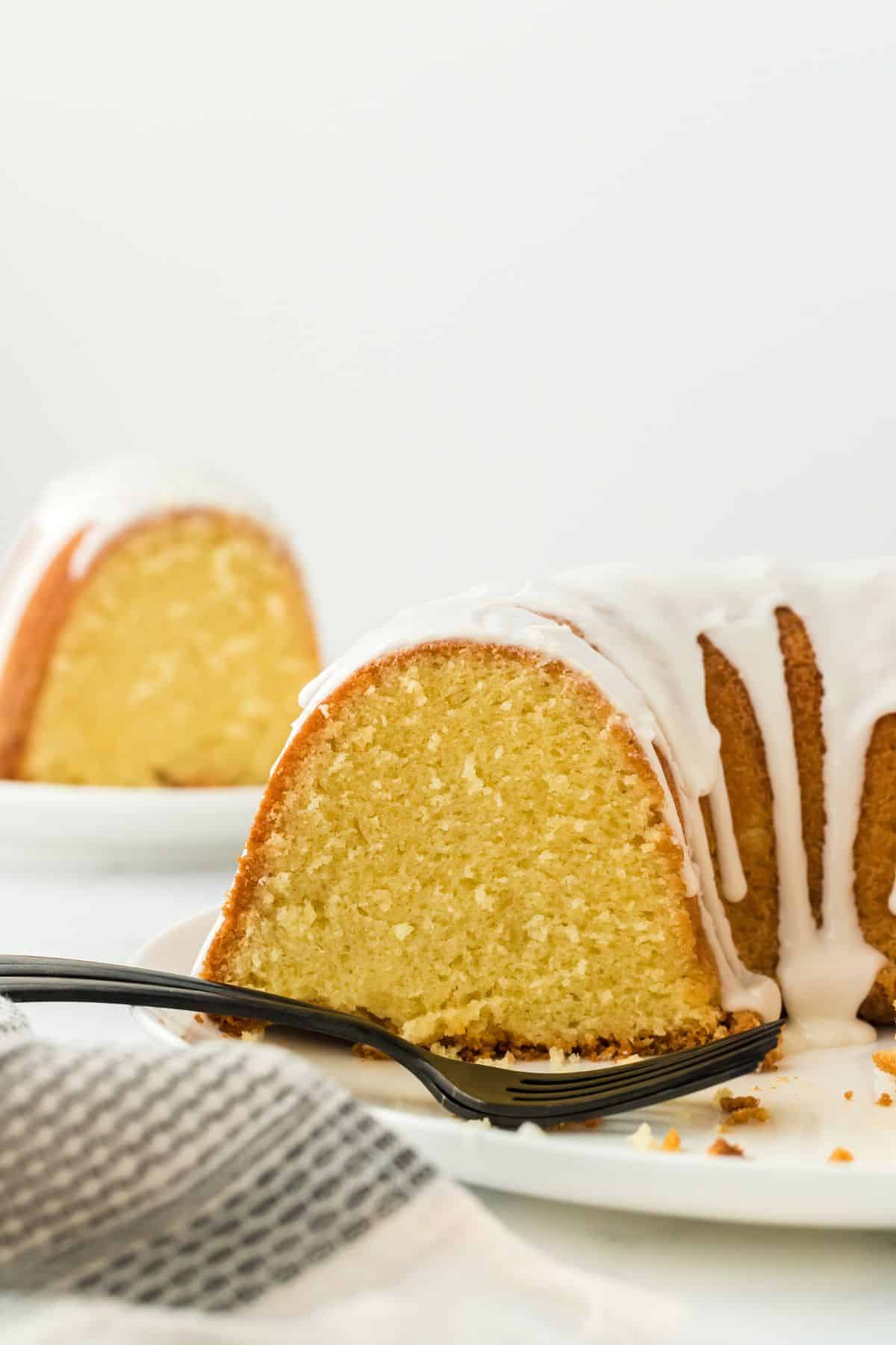 Closeup of a thick slice of five flavor pound cake with white icing, accompanied by a black fork and a patterned cloth and more cake in the background