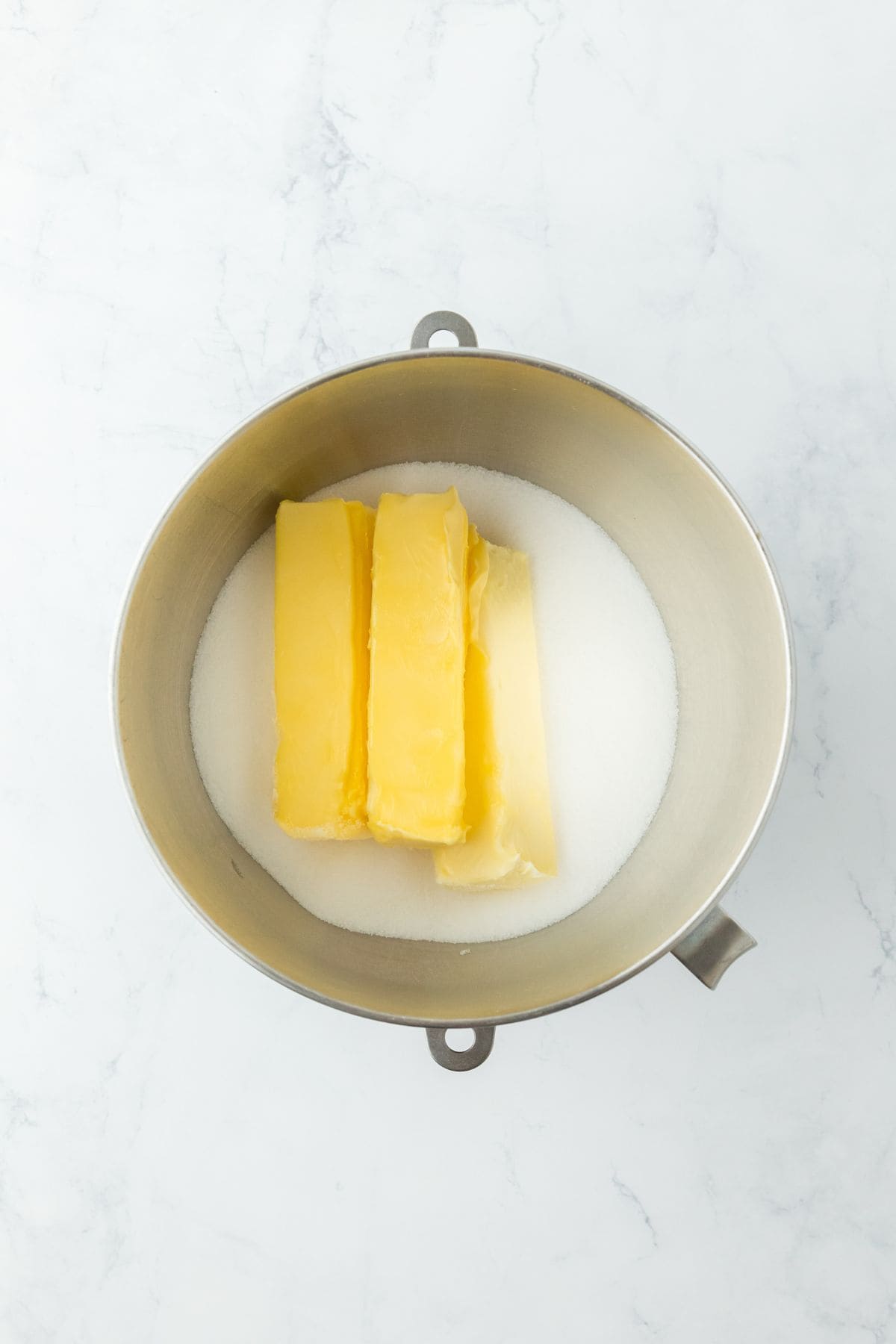 A mixing bowl with sticks of butter and sugar at the bottom, ready to be creamed together