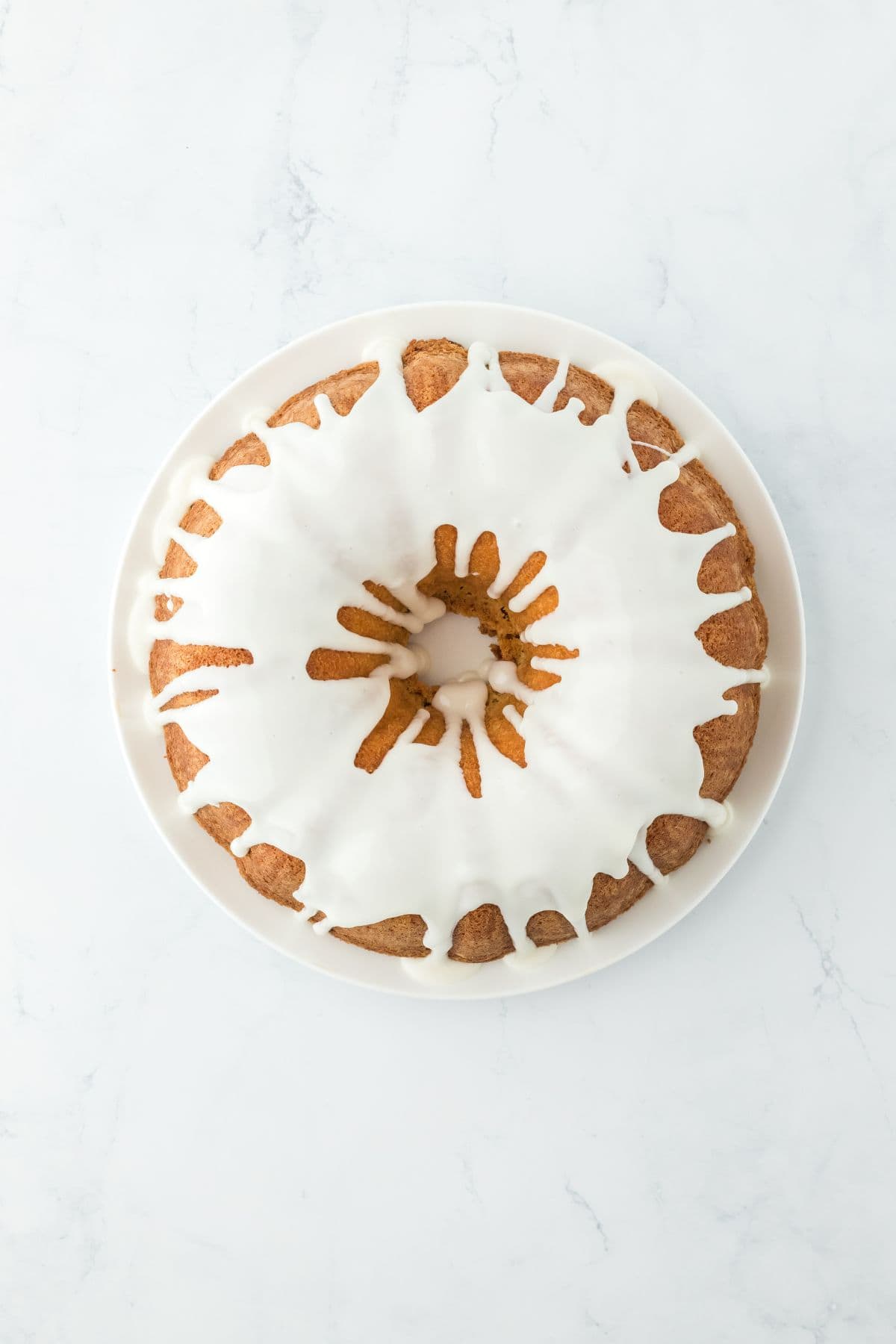 An overhead view of a five flavor pound cake with white icing drizzled over it, fully coated on a white plate