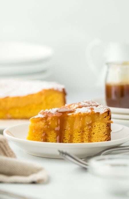 A gooey brown butter cake slice with a drizzle of caramel sauce, showcasing the rich, golden interior with the rest of the cake and a caramel jar in the background