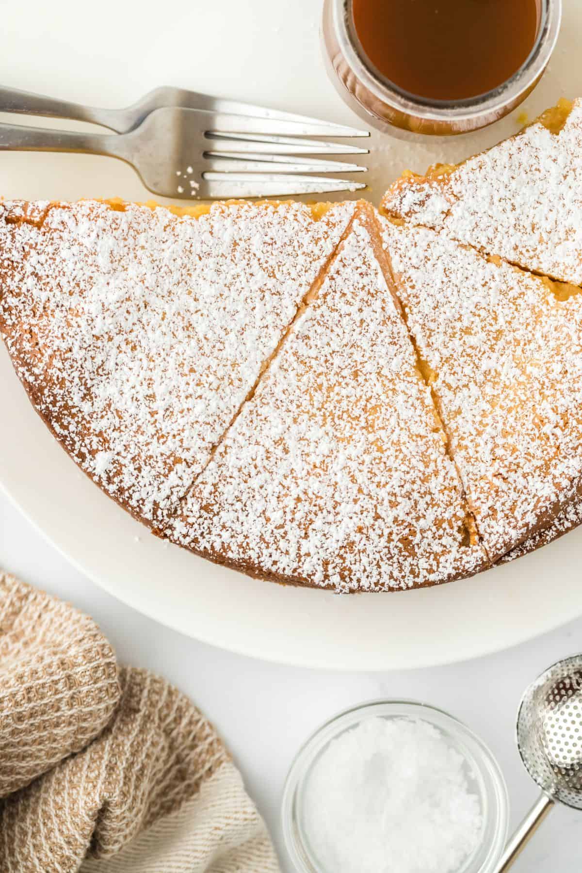 Overhead shot of gooey brown butter cake sliced on a plate, dusted with powdered sugar, with forks and caramel sauce jar on the side