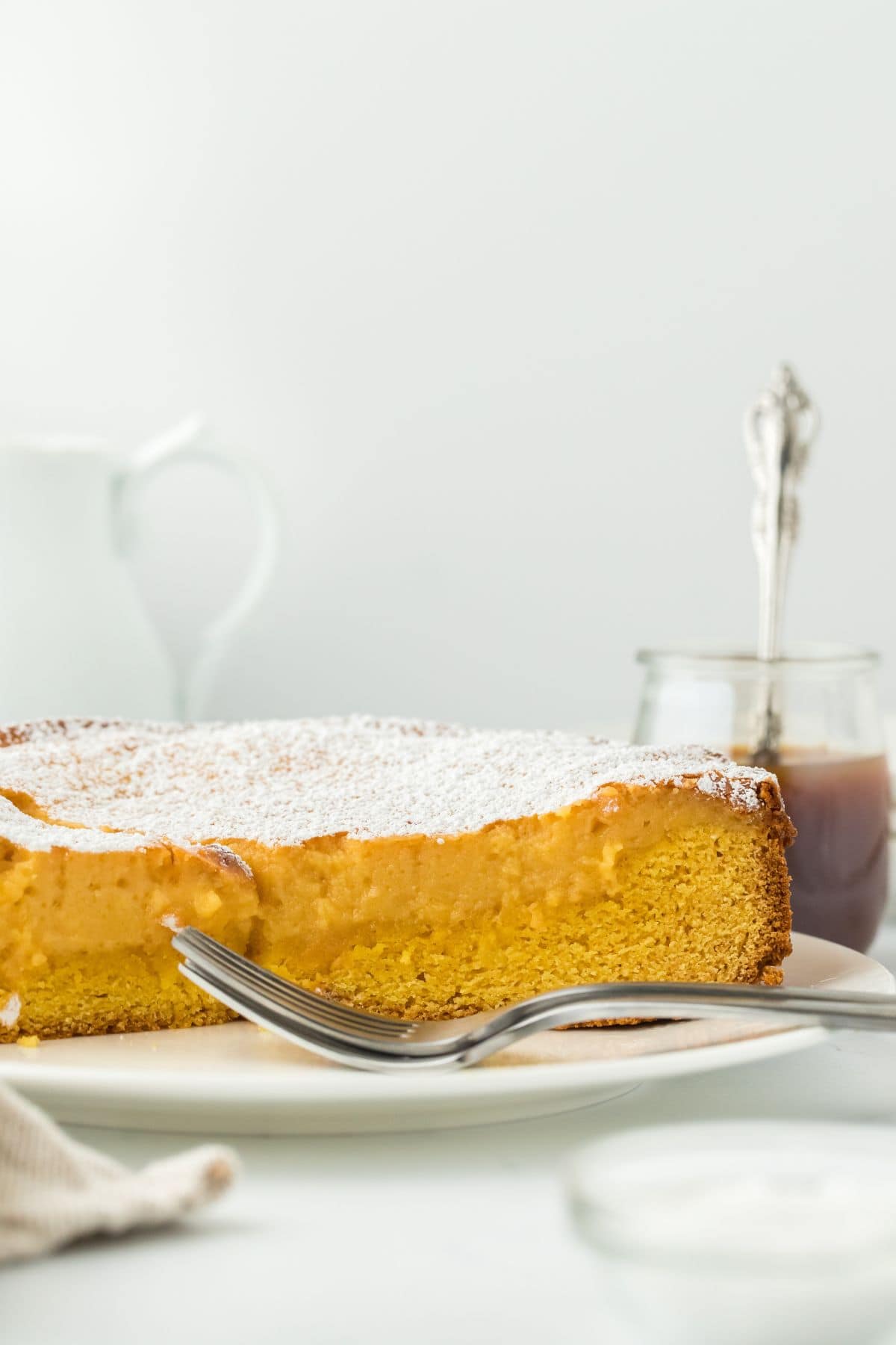 A slice of gooey brown butter cake on a plate, with a fork next to it and a jar of caramel in the background