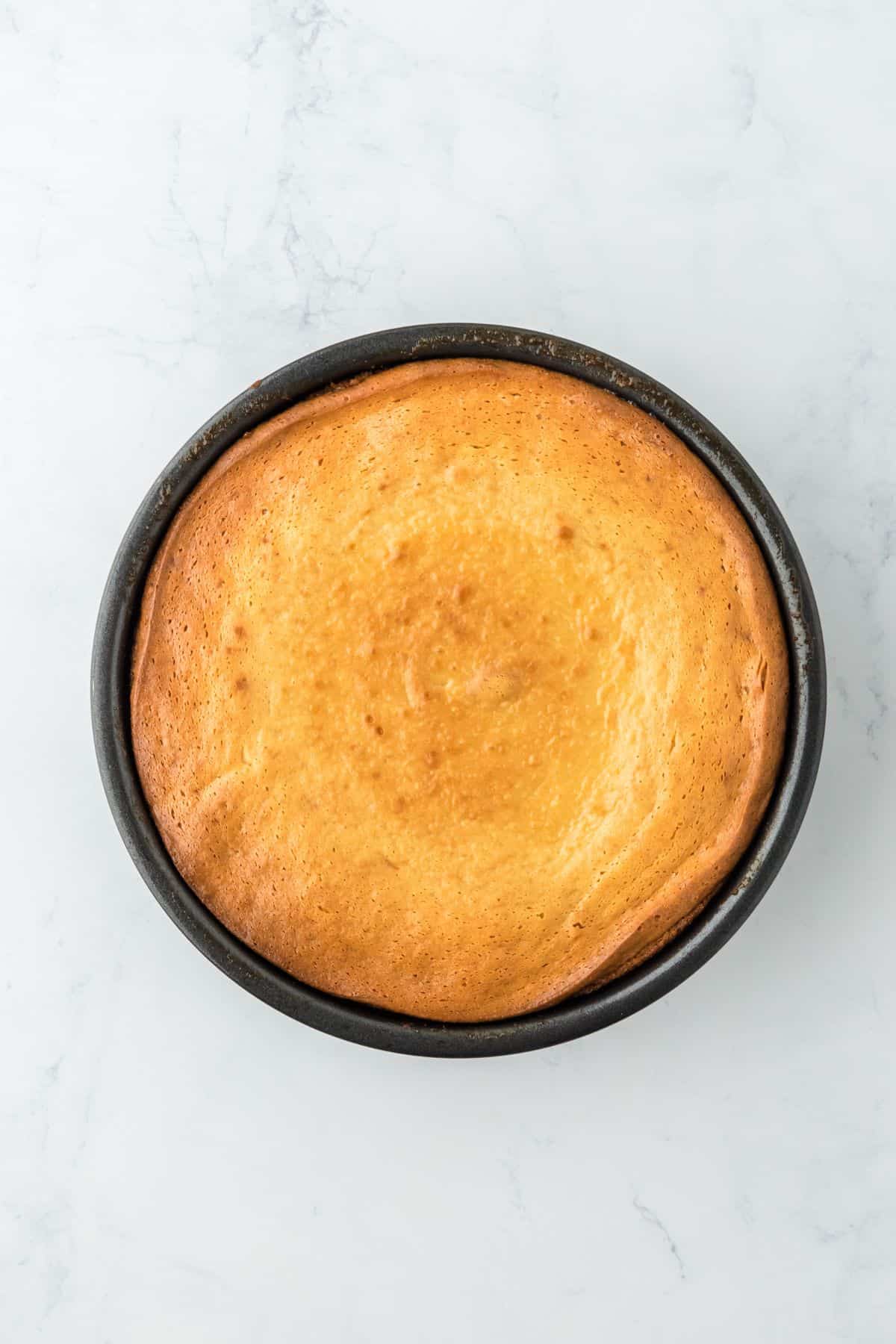 A browned, baked cake in a round pan, showing a lightly cracked, golden-brown surface