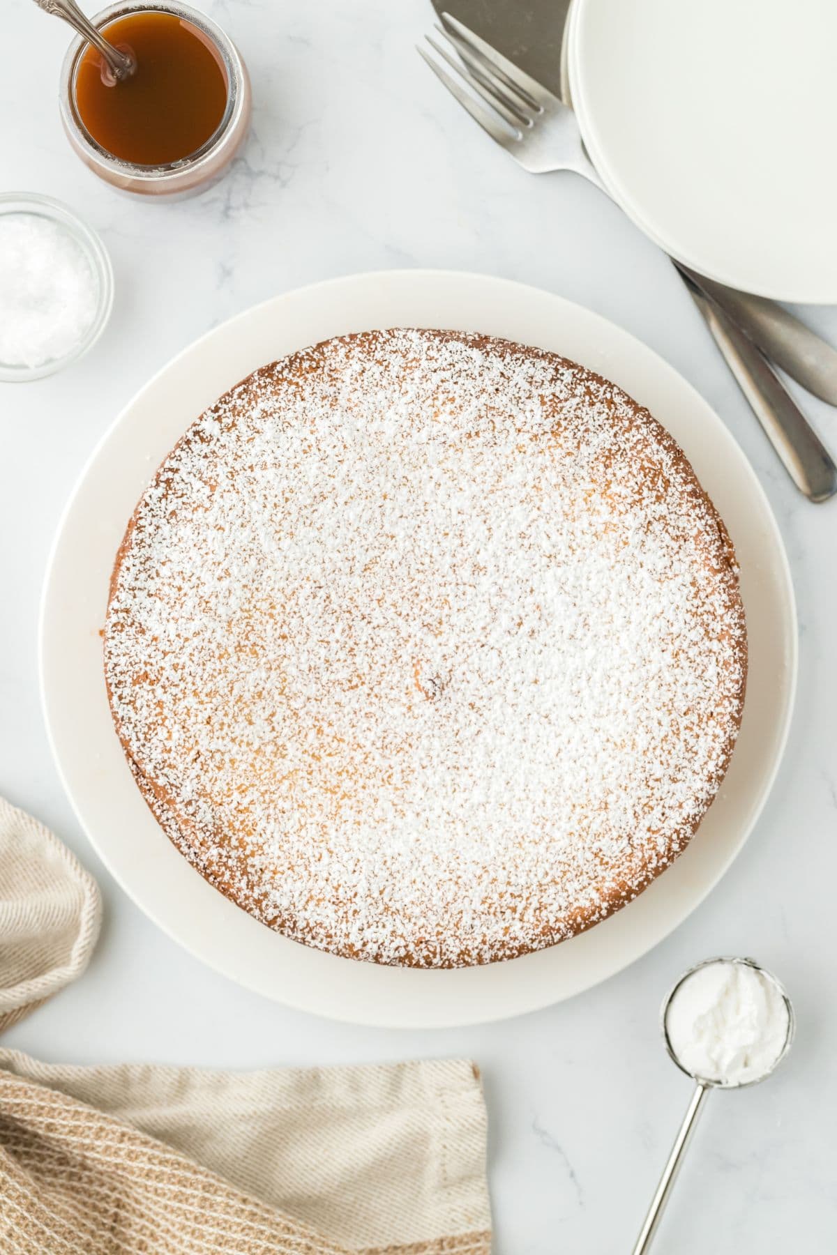 A powdered sugar-dusted gooey brown butter cake placed on a white plate, with a jar of caramel sauce and utensils nearby