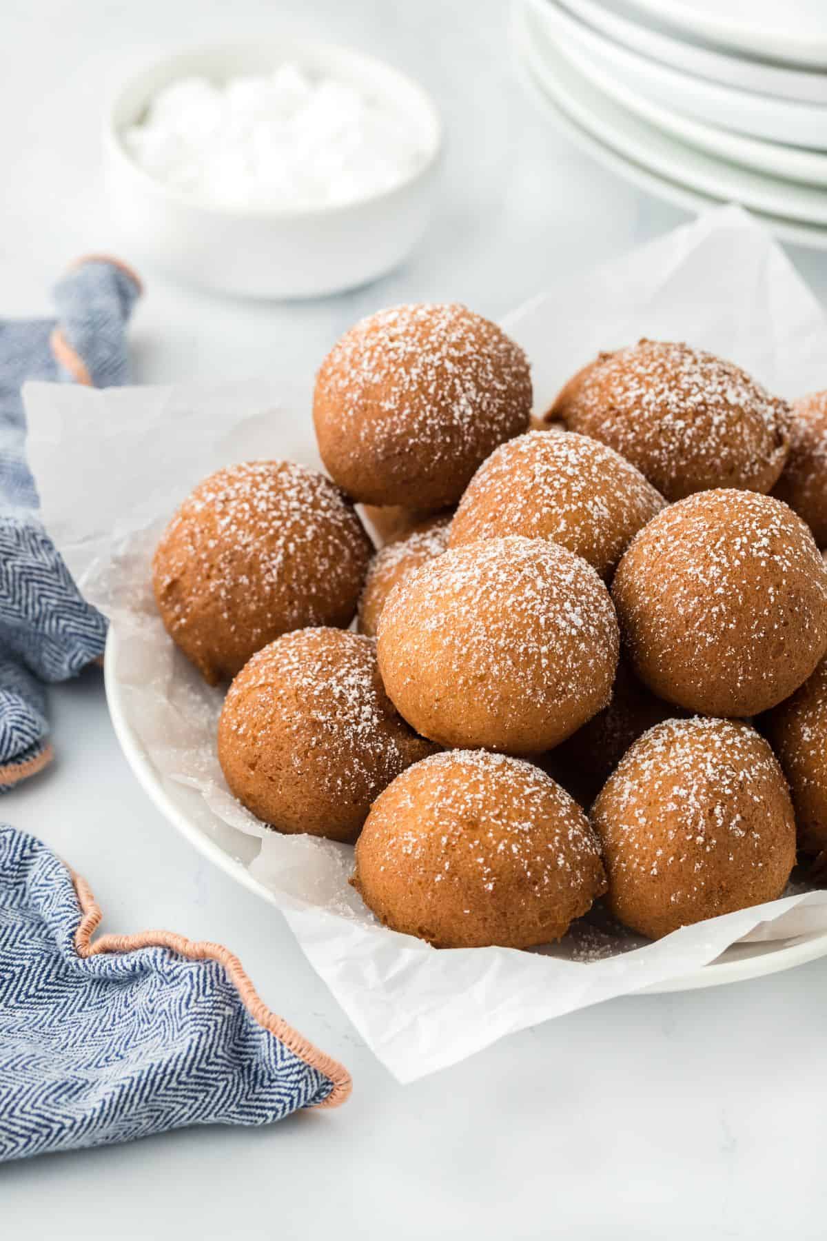 A plate of pound cake beignets dusted with powdered sugar, with a blue cloth napkin and a bowl of powdered sugar nearby