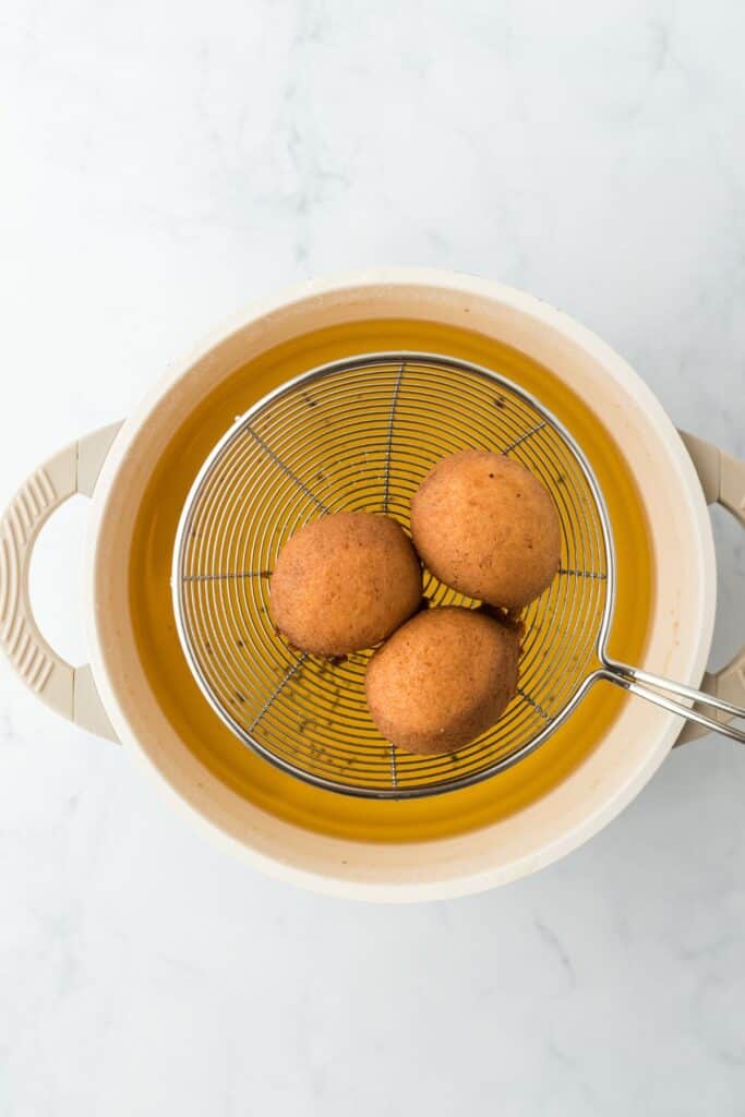 Three round, golden-brown beignets freshly fried in a round oil strainer above a pot of oil