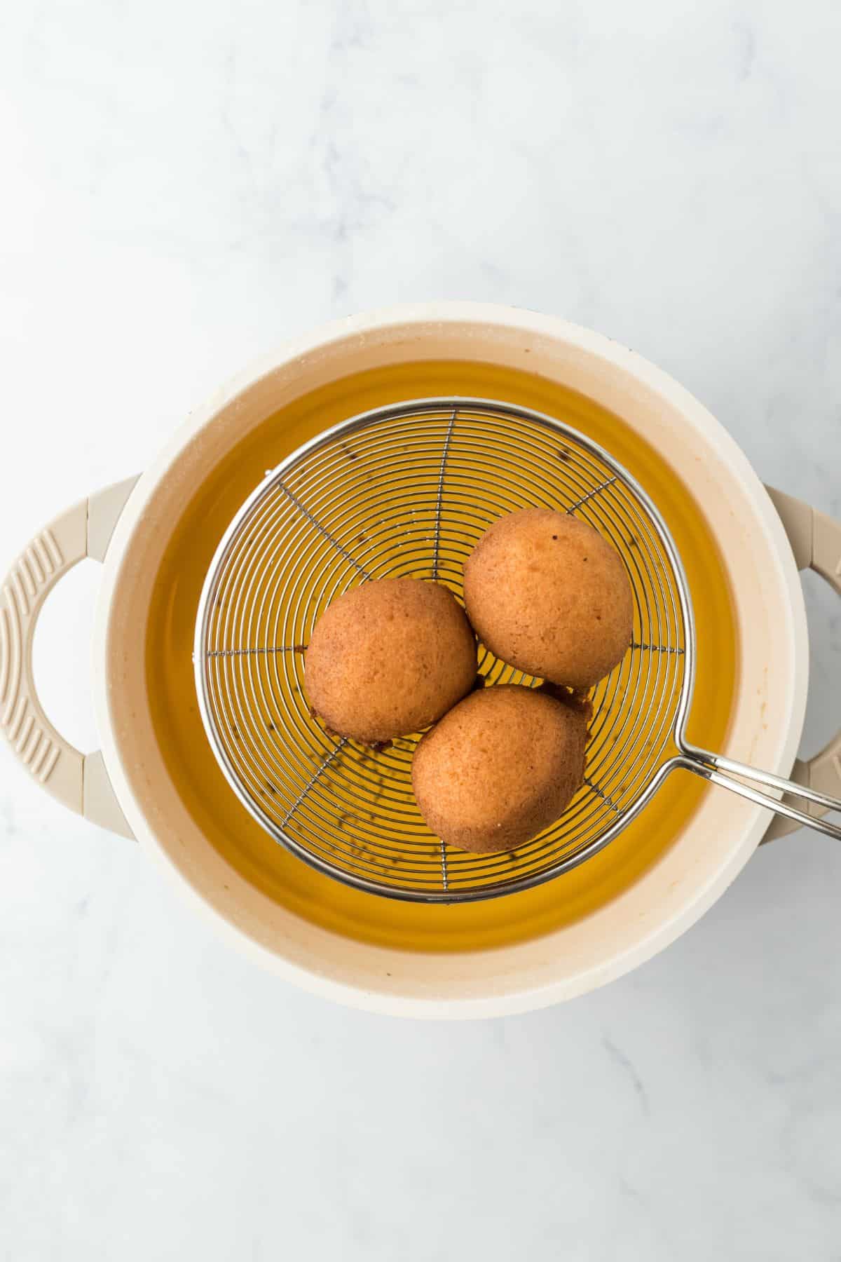 Three round, golden-brown beignets freshly fried in a round oil strainer above a pot of oil