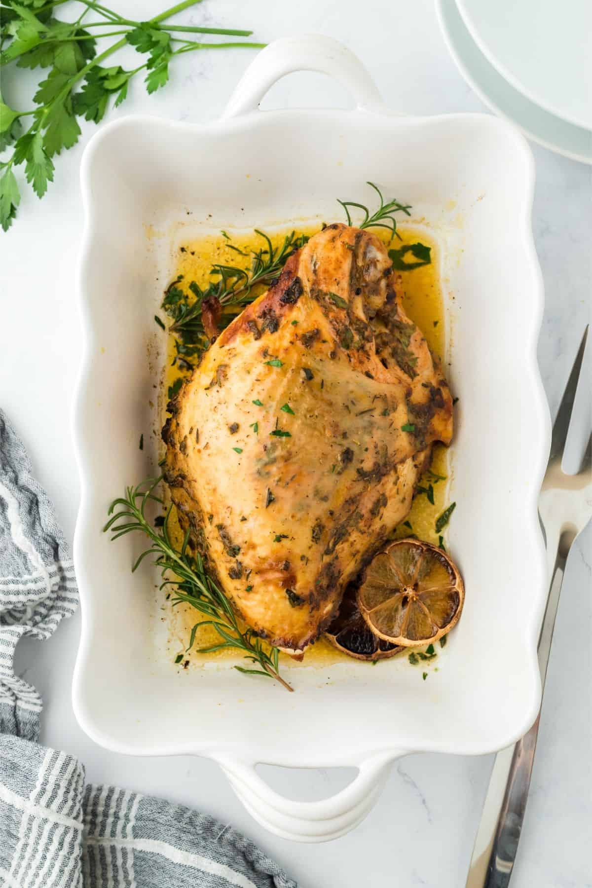 Overhead shot of a golden-brown roasted turkey breast seasoned with herbs, garnished with lemon slices and fresh rosemary in a white baking dish