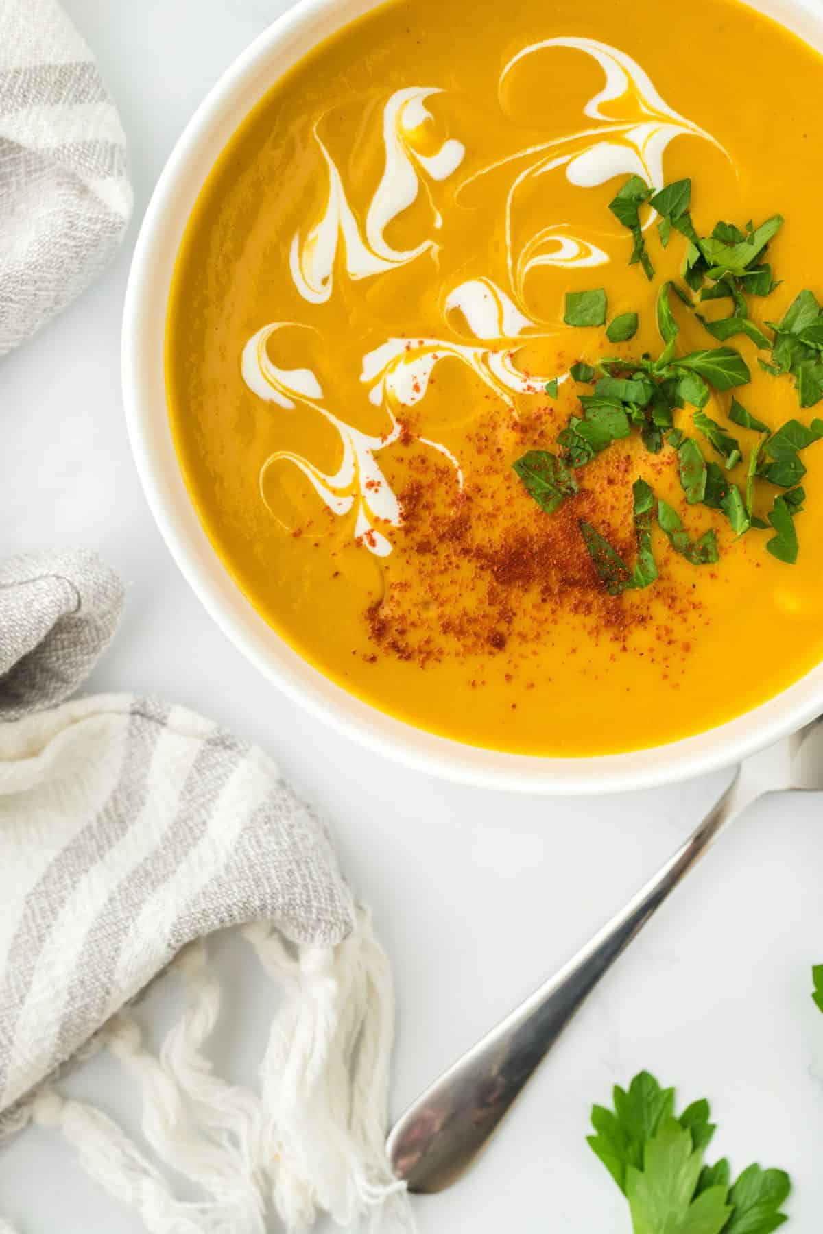 Overhead shot of sweet potato soup in a white bowl with swirls of cream and garnished with parsley and paprika, next to a spoon and a napkin