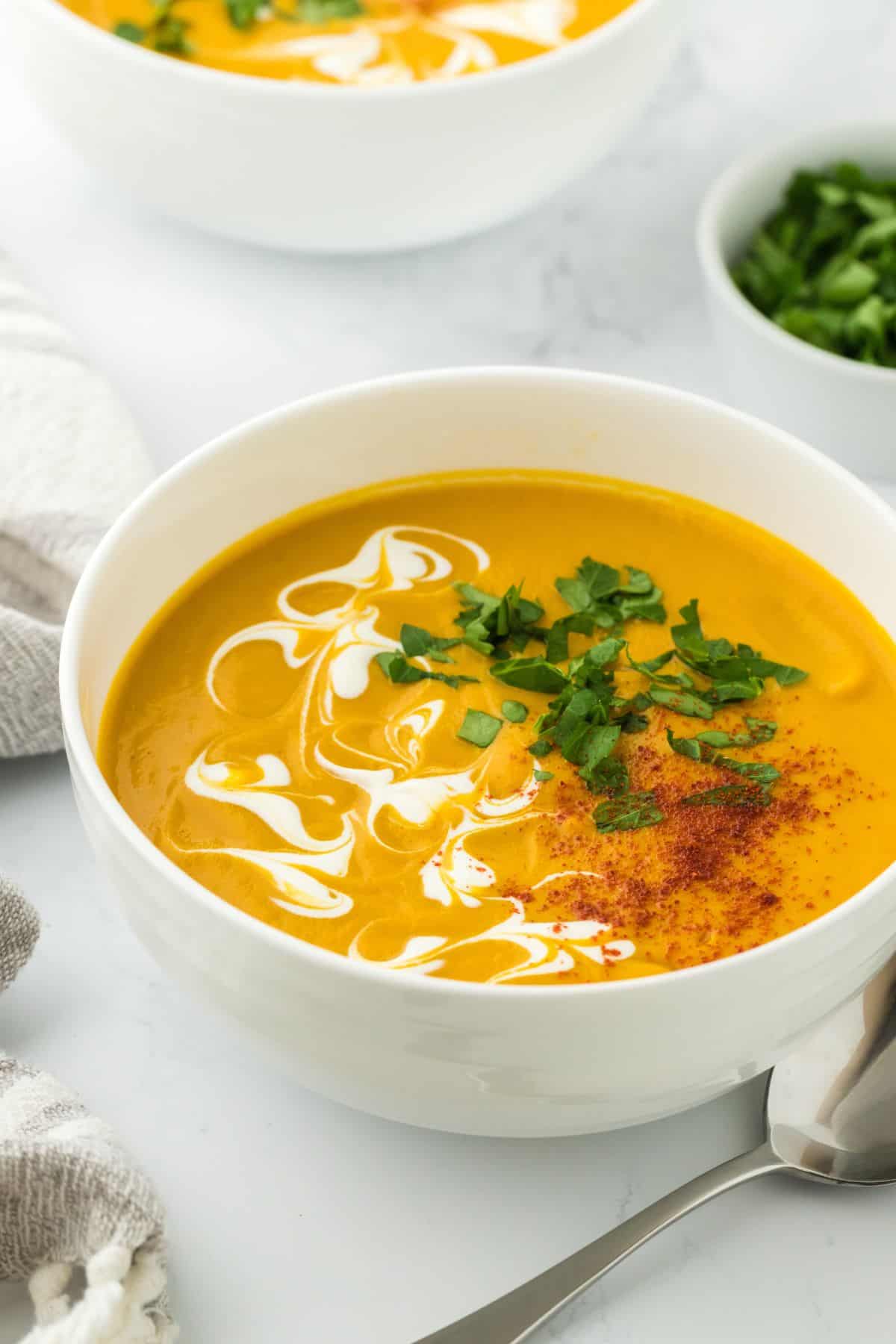 Sweet potato soup in a white bowl, garnished with cream swirls, chopped parsley, and paprika, with a side of extra parsley in a small dish