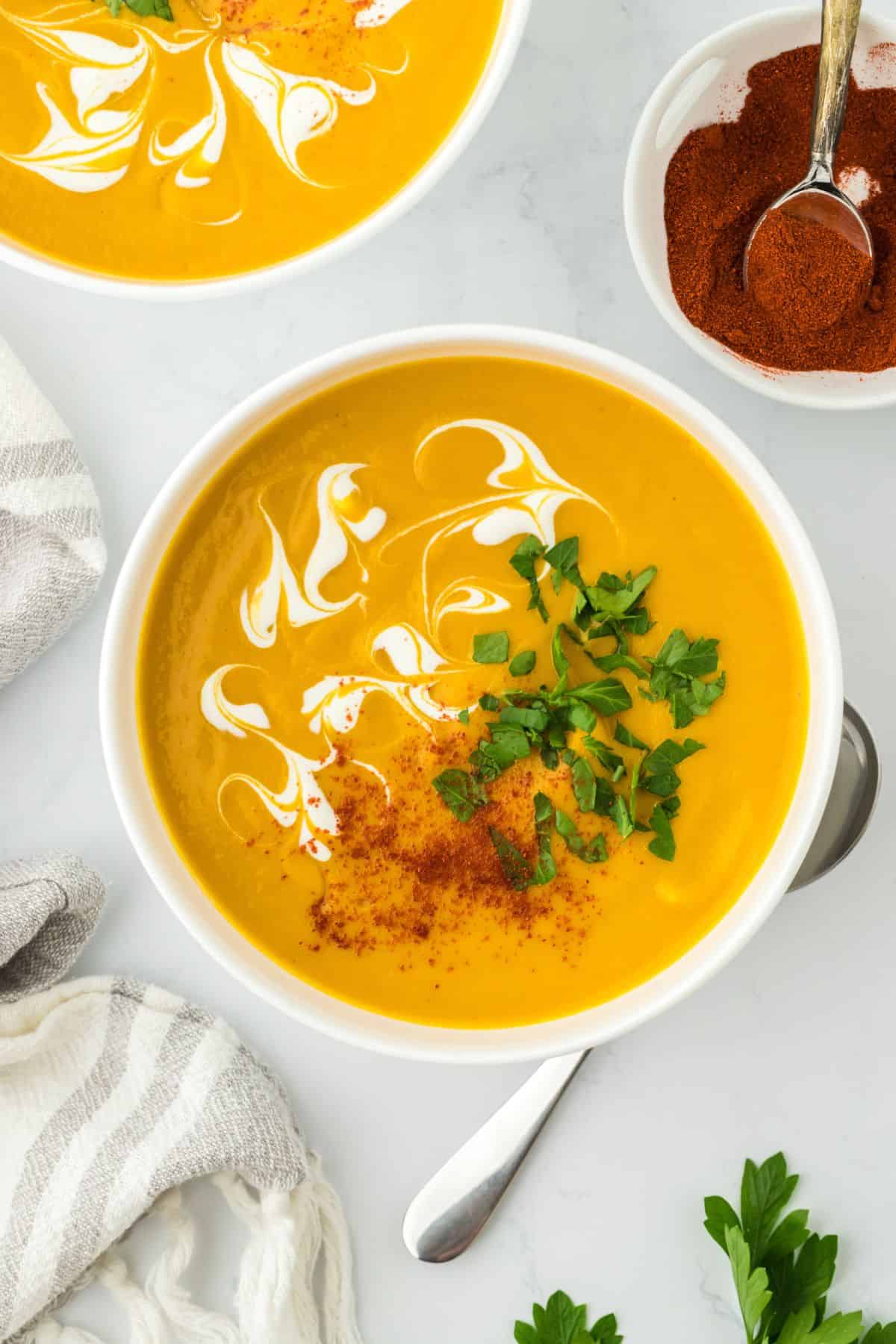 Overhead shot of a bowl of creamy sweet potato soup topped with a swirl of cream, a sprinkle of paprika, and chopped parsley for garnish, with a cloth napkin, more soup and bowl of spices in the background