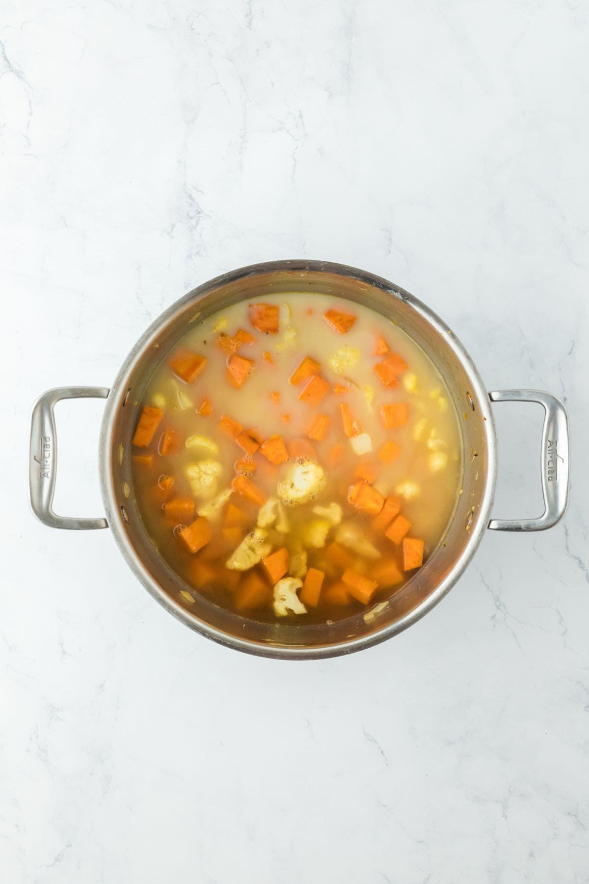 A pot filled with simmering vegetables in a broth, including sweet potatoes, onions, and cauliflower, beginning to soften